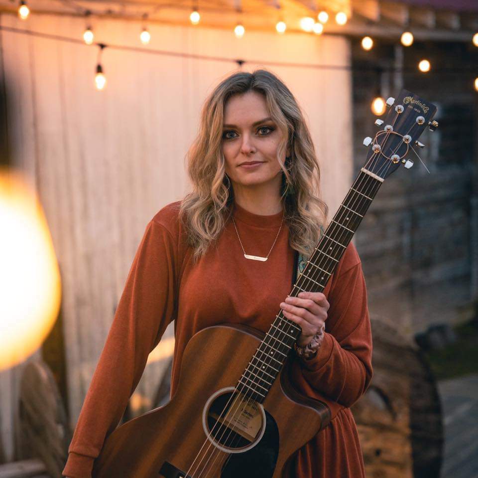 A person with long, wavy hair is standing outside, holding an acoustic guitar. They are wearing a rust-colored long-sleeve dress and smiling softly at the camera. String lights are hanging in the background, creating a warm and cozy atmosphere.