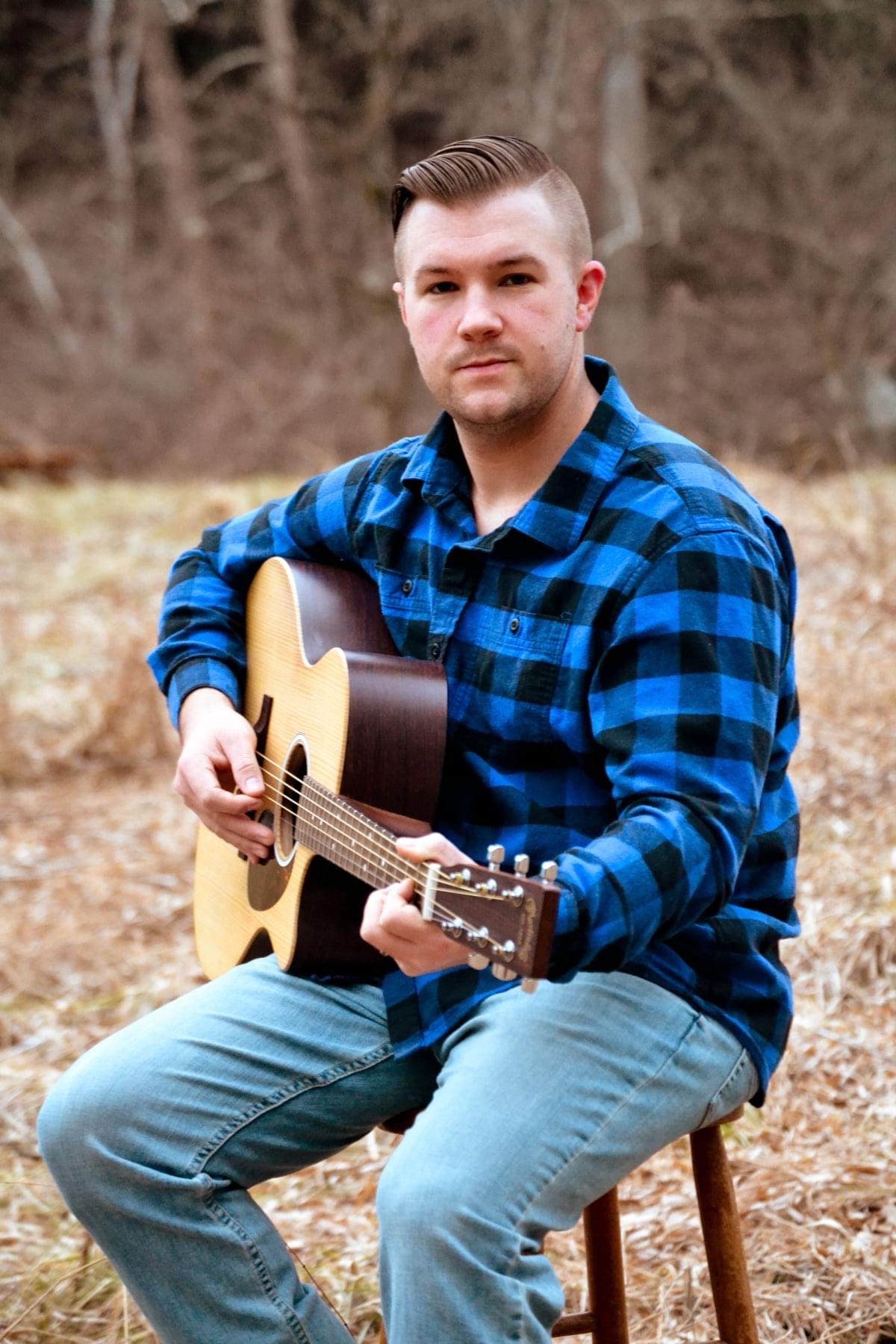 A person with short hair in a blue and black plaid shirt and jeans sits on a stool outdoors, playing an acoustic guitar. The background features dry grass and trees, suggesting it is autumn or winter. The person looks directly at the camera.