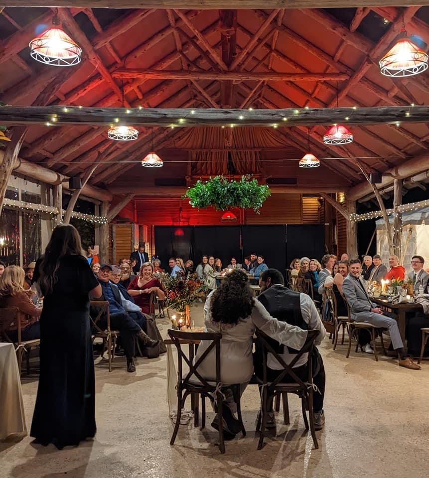 A couple sits closely together at the front of a decorated rustic venue with wooden beams and hanging lights. They are facing a large gathering of seated guests, who are attentively watching a standing woman speak into a microphone. The atmosphere is warm and lively.