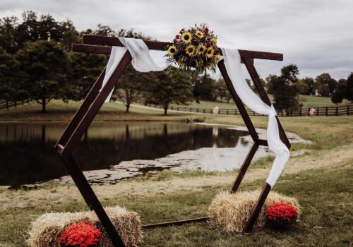 A hexagonal wedding arch stands by a pond, decorated with sunflowers, white draped fabric, and surrounded by bundles of hay. Red potted flowers are placed on the ground beside the hay. Trees and cloudy sky are seen in the background. Image
