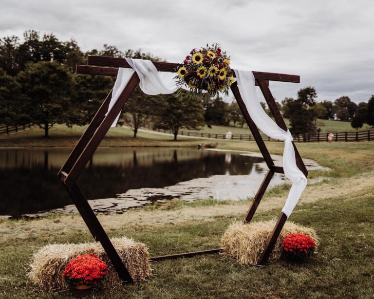 A hexagonal wedding arch stands by a pond, decorated with sunflowers, white draped fabric, and surrounded by bundles of hay. Red potted flowers are placed on the ground beside the hay. Trees and cloudy sky are seen in the background.
