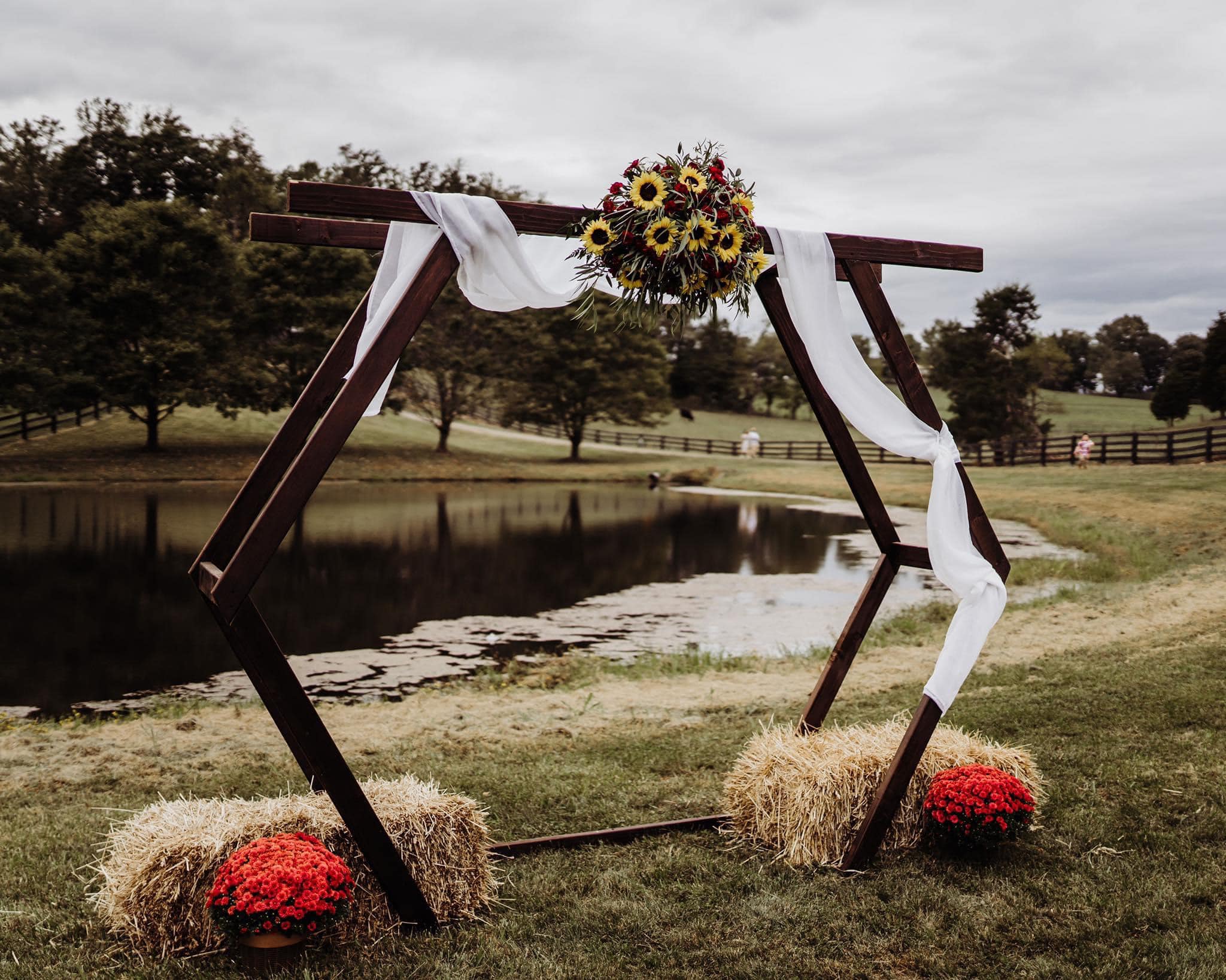 A hexagonal wedding arch stands by a pond, decorated with sunflowers, white draped fabric, and surrounded by bundles of hay. Red potted flowers are placed on the ground beside the hay. Trees and cloudy sky are seen in the background.