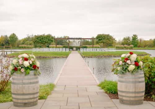 A picturesque outdoor wedding setup featuring a long wooden aisle leading to an archway adorned with flowers. White chairs are neatly arranged on both sides of the aisle, flanked by wine barrels at the front, with a scenic view of trees and water surrounding the area. Image