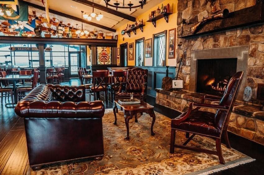 A cozy room with a stone fireplace and leather seating. The room features a plush rug, a wooden coffee table, and ornate decor. Wooden shelves display bottles and other items above the seating area. A chandelier hangs from the ceiling, and windows line one wall.