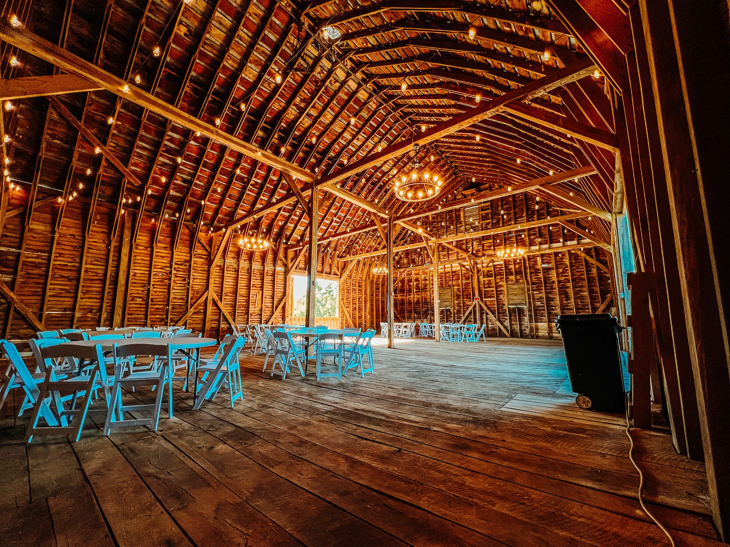 A spacious, rustic barn venue with wooden floors and walls. Bright string lights and chandeliers hang from the vaulted ceiling. Round tables with blue chairs are arranged throughout the space, suggesting it's set up for an event or gathering.