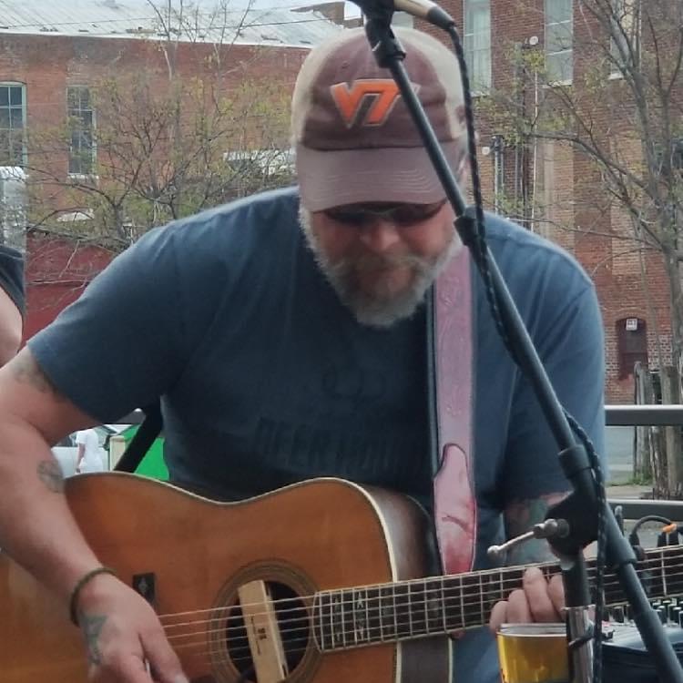 A man wearing a baseball cap and sunglasses is playing an acoustic guitar and singing into a microphone. He is sitting outdoors, with a building and some trees visible in the background. A glass of beer is placed in front of him.