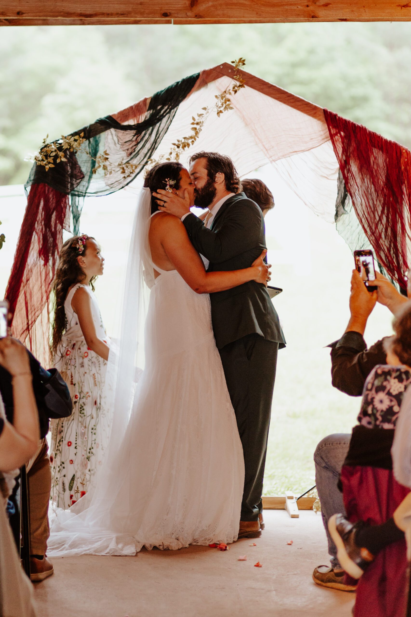 A bride and groom share a kiss under a decorated arch at a rustic wedding venue in Virginia. The bride is wearing a white dress and veil, and the groom is in a dark suit. A child in a floral dress stands nearby, while guests capture the magical moment on their phones.