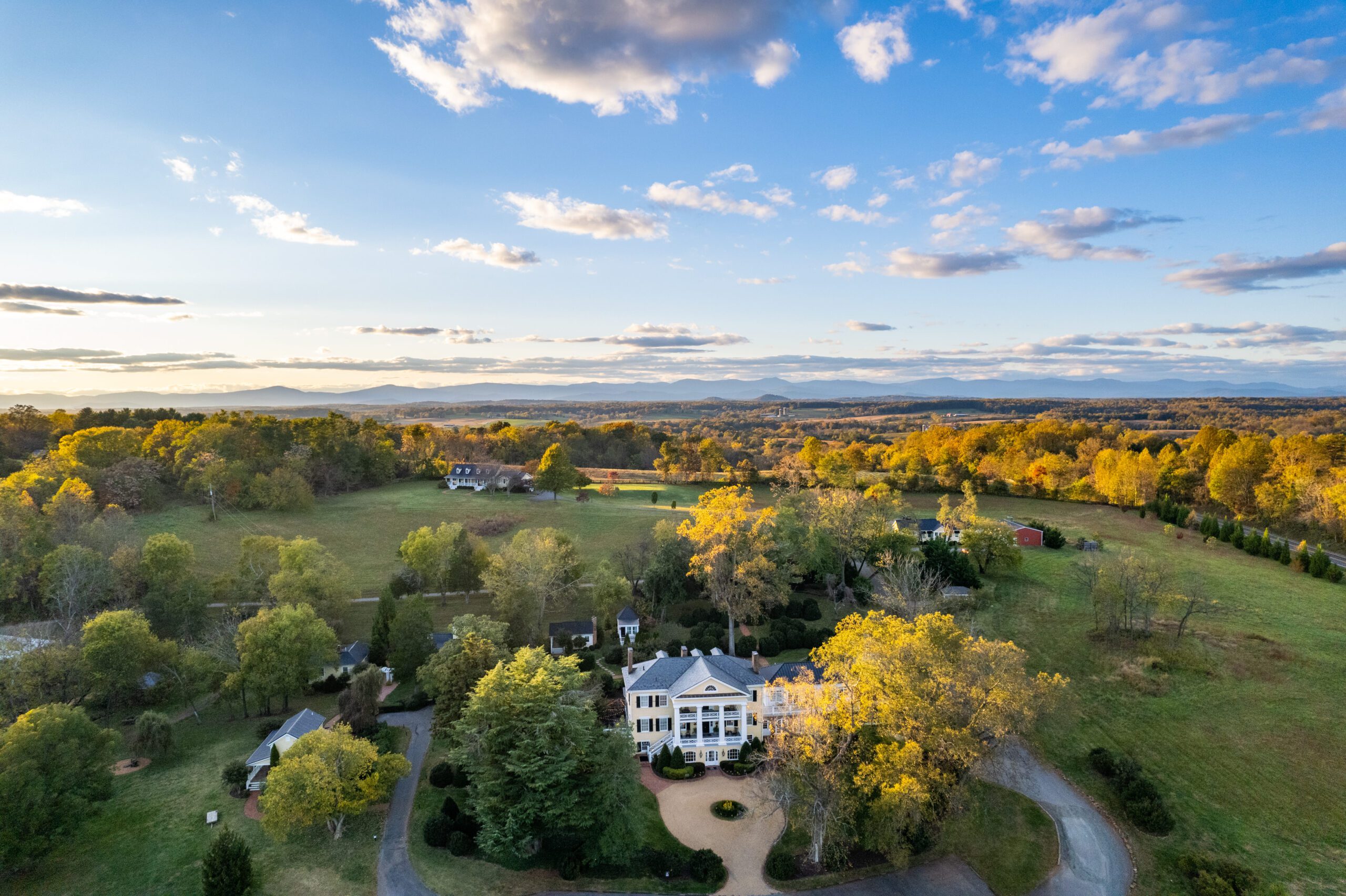 Aerial view of a large white colonial-style house surrounded by trees with changing leaves. The property includes a winding driveway and open fields. In the distance, there are rolling hills and mountains under a blue sky with scattered clouds.