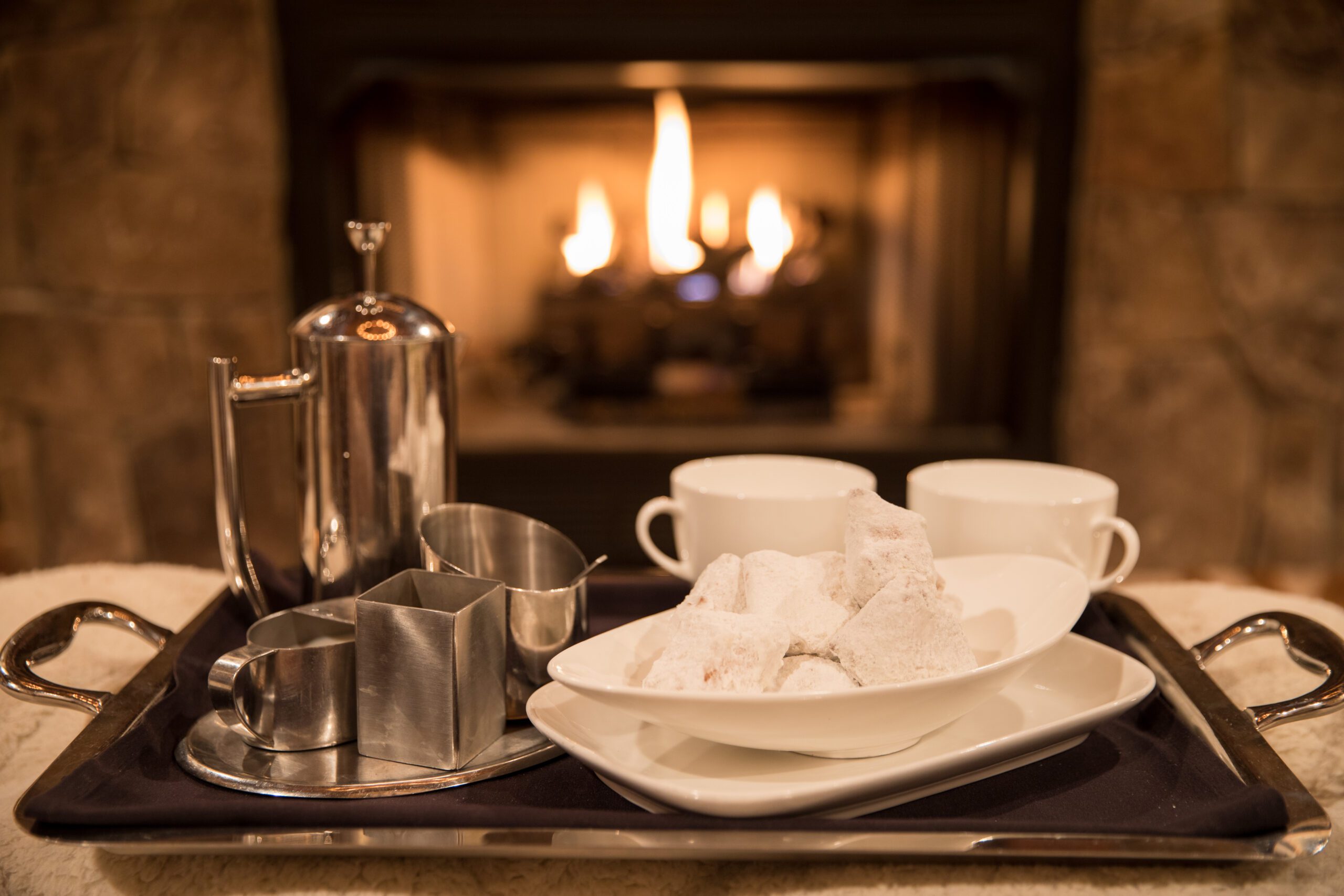 A cozy scene features a tray with a shiny silver coffee pot, milk jug, sugar bowl, and two white cups, placed in front of a lit fireplace. A bowl of powdered sugar-coated treats is also on the tray, adding a touch of warmth and comfort.