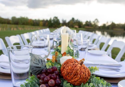 A long outdoor dining table is elegantly set with white tablecloths, plates, glasses, and cutlery. The table features an autumn-themed centerpiece with grapes, pumpkins, eucalyptus, and candles. A 
