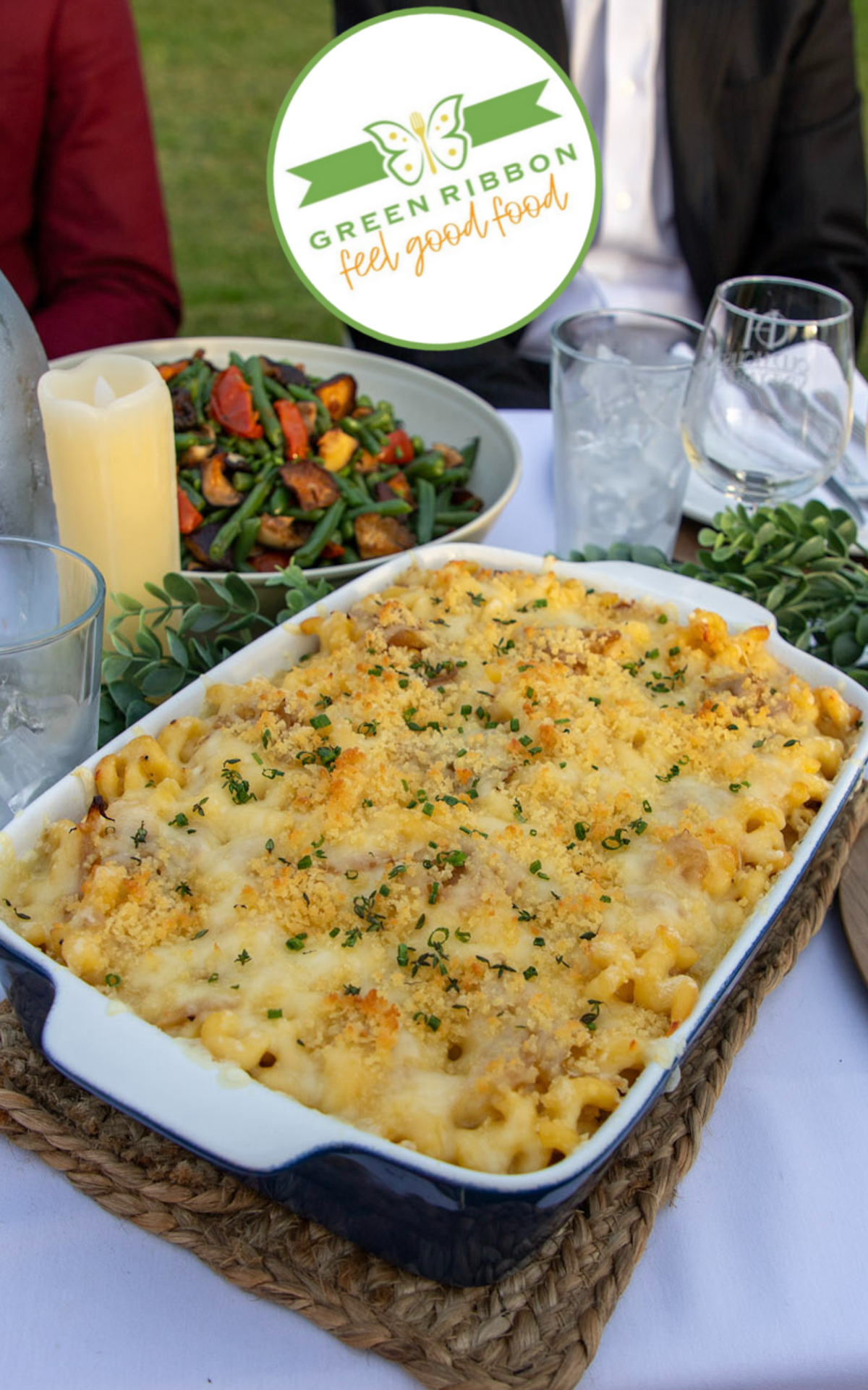 A casserole dish filled with cheesy pasta is placed on a woven placemat on an outdoor table. Next to it are a candle, glasses of water, and a plate of colorful roasted vegetables. The background has a blurred greenery. A logo above reads 
