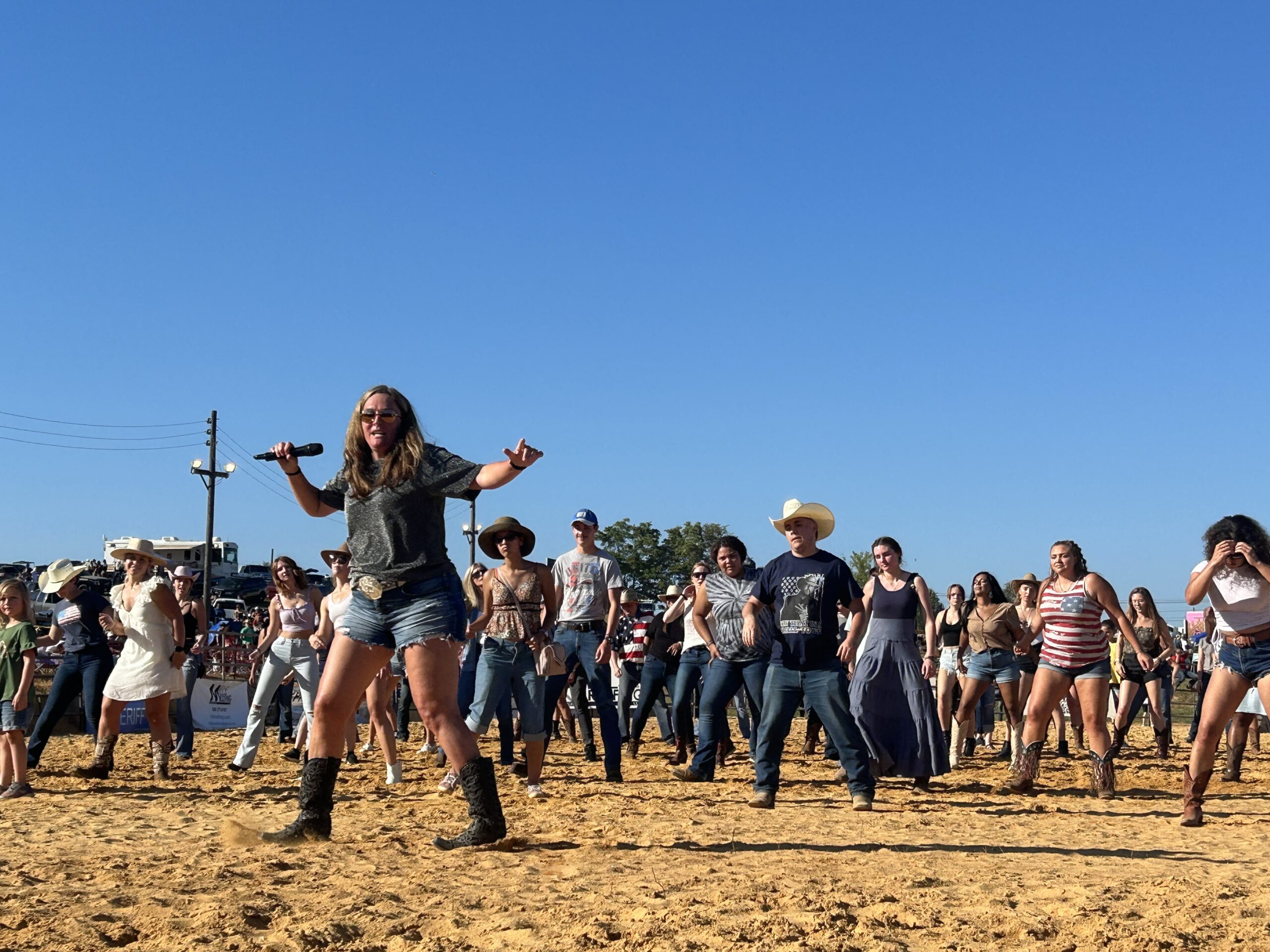 A group of people participating in a line dance on a sandy ground. The crowd includes men and women in casual attire, many wearing cowboy hats and boots. The sky is clear and blue, indicating a sunny day. The leader is in the foreground guiding the dance.