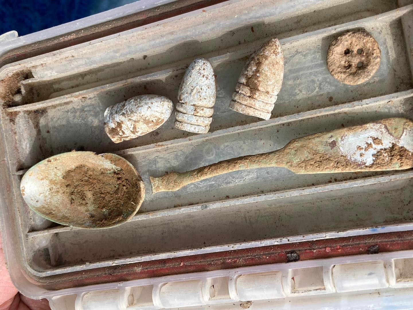 A weathered compartmentalized box containing three old bullets, a tarnished spoon, and a worn button. The items are covered in dirt and look aged, suggesting they may be historical artifacts.