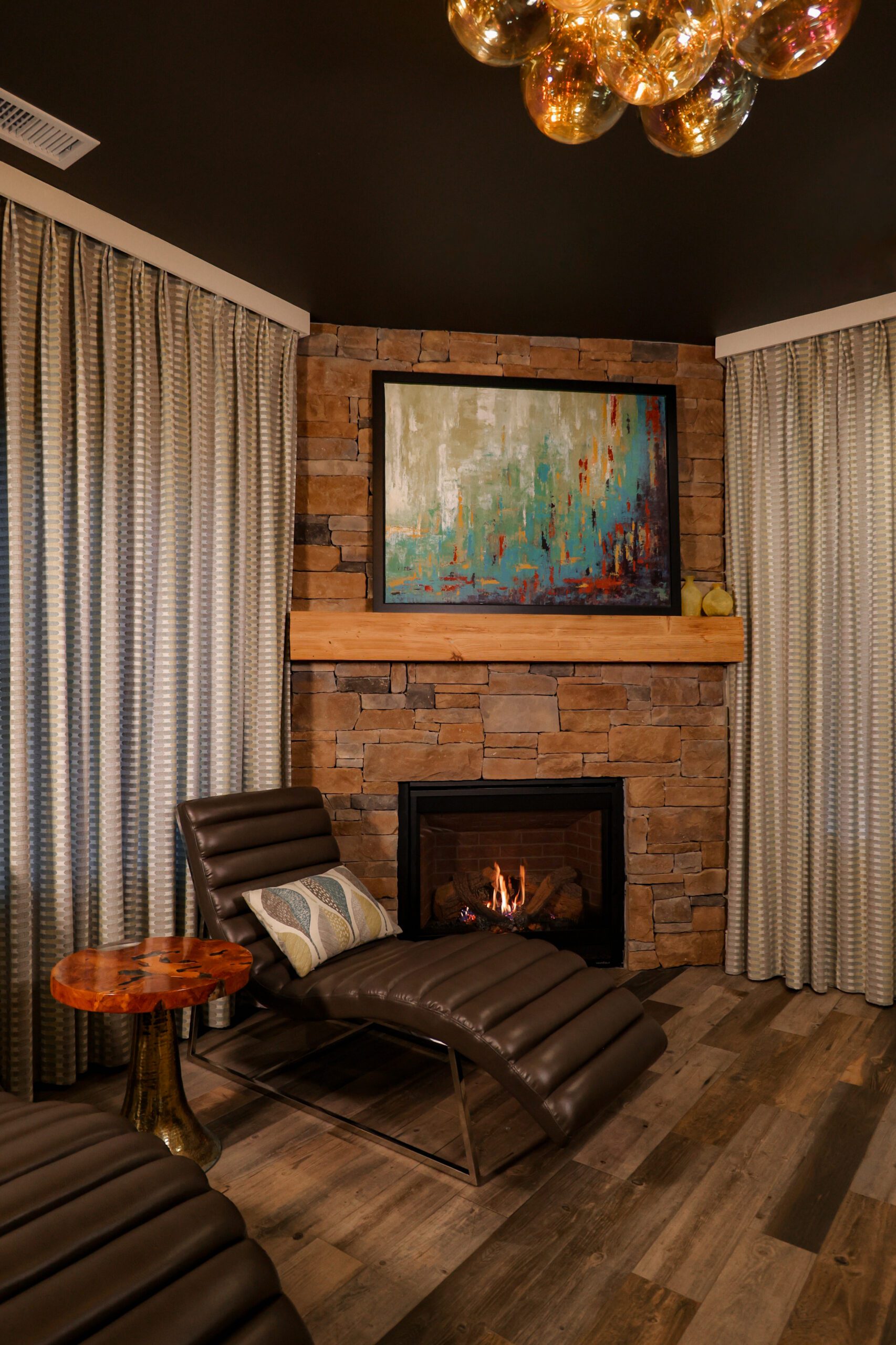 A cozy room features a stone fireplace with a lit fire, above which hangs an abstract painting in blue, green, and orange tones. Two brown leather lounge chairs with pillows face the fireplace, accompanied by a small wooden side table. Beige curtains frame the scene.