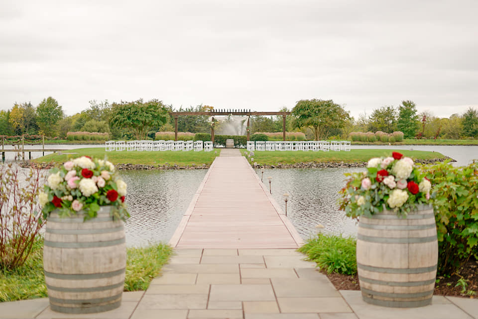 A wooden walkway extends over a pond toward an outdoor wedding venue in Virginia, with rows of white chairs facing a floral arch. The walkway is flanked by two large barrels adorned with bouquets of white and red flowers. Greenery and trees are visible in the background.