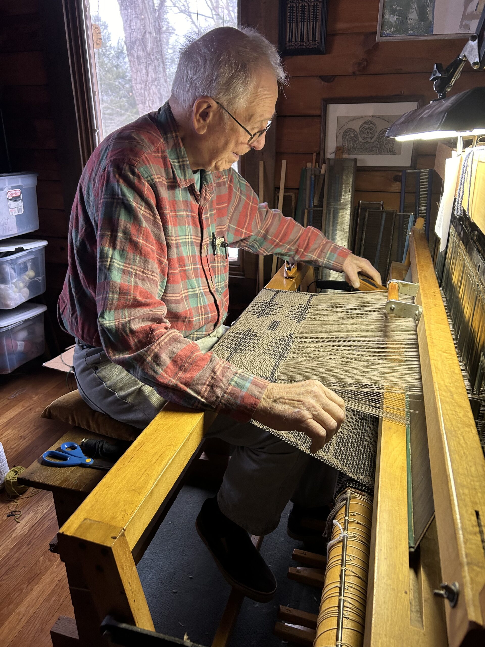 An elderly man wearing a red plaid shirt and glasses is seated at a large, wooden weaving loom. He's focusing intently on weaving a complex pattern into a fabric. The room is rustic with wooden walls and various weaving tools around him.
