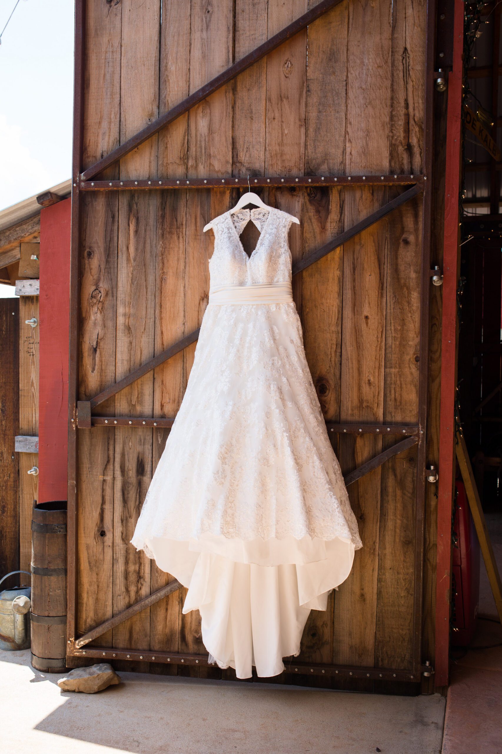 A white lace wedding dress with a sleeveless V-neck design hangs on a wooden barn door, captured beautifully by a Virginia Wedding Photographer. The dress features a fitted bodice, satin waistband, and flowing A-line skirt with multiple fabric layers visible at the bottom. The door is secured with metal brackets.