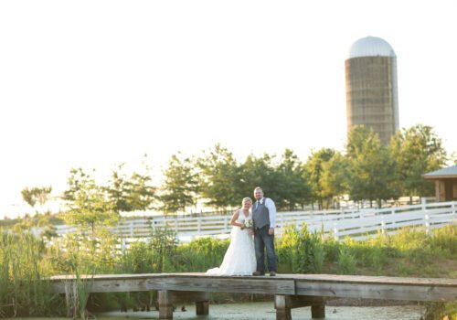 A bride and groom pose together on a wooden footbridge amidst a lush, green field. The bride is in a white wedding dress, and the groom is in a vest with rolled-up sleeves. Captured by a top Virginia Wedding Photographer, they stand before a white fence, trees, and a tall silo under a bright sky. Image