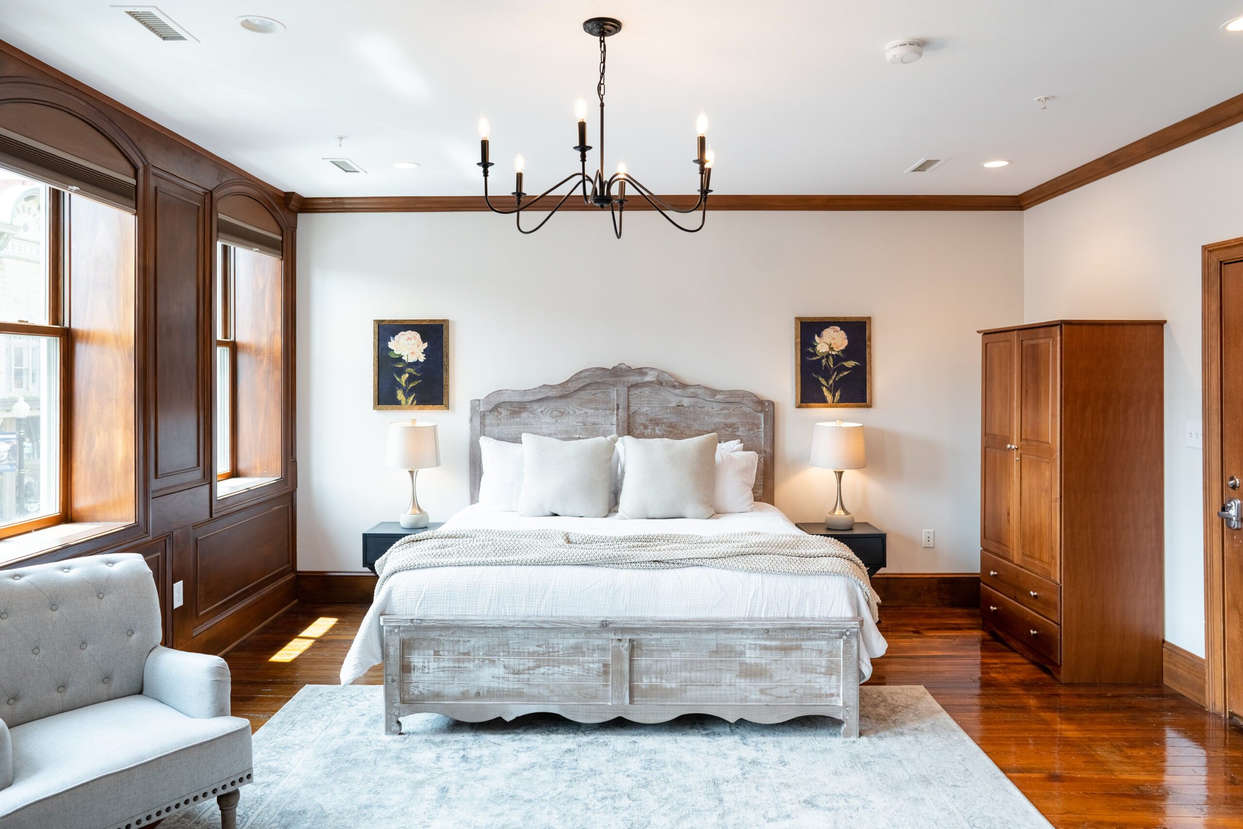 A cozy bedroom ideal for a weekend getaway, featuring a large bed with a wood headboard, white pillows, and blankets. It is flanked by wooden nightstands with lamps. The room includes a wooden wardrobe, a gray armchair, hardwood floors, and an overhead chandelier. Two paintings hang above the nightstands.