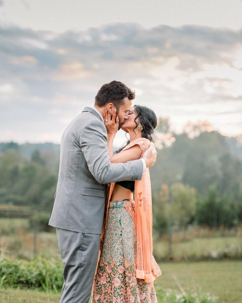 A couple shares a kiss outdoors at sunset. The man wears a light gray suit, and the woman wears a vibrant, floral-embroidered traditional dress with a flowing shawl. They embrace each other lovingly with lush greenery and scenic hills in the distance, highlighting one of the stunning wedding venues in Virginia.