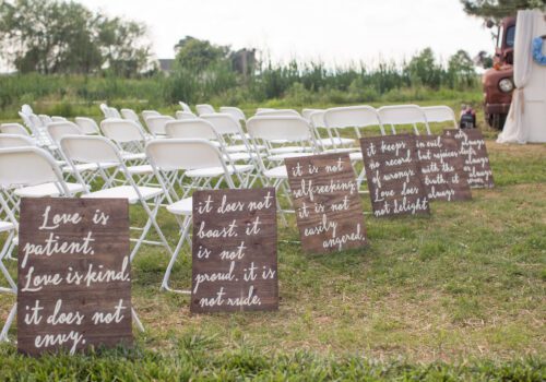 Rows of white folding chairs are arranged outdoors on a grassy lawn for a wedding ceremony at one of the most charming wedding venues in Virginia. Wooden signs with handwritten quotes about love are placed along the aisle, with tall grasses and trees in the background. An old-fashioned truck is visible to the right. Image