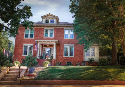 A large, two-story red brick house with multiple windows and a small front porch sets the perfect scene for a weekend getaway. An American flag hangs beside the entrance. The house is surrounded by greenery, with a few trees and bushes in the well-kept front lawn. A sunny sky is visible in the background.