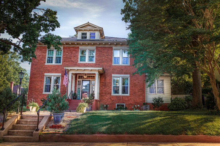 A large, two-story red brick house with multiple windows and a small front porch sets the perfect scene for a weekend getaway. An American flag hangs beside the entrance. The house is surrounded by greenery, with a few trees and bushes in the well-kept front lawn. A sunny sky is visible in the background.