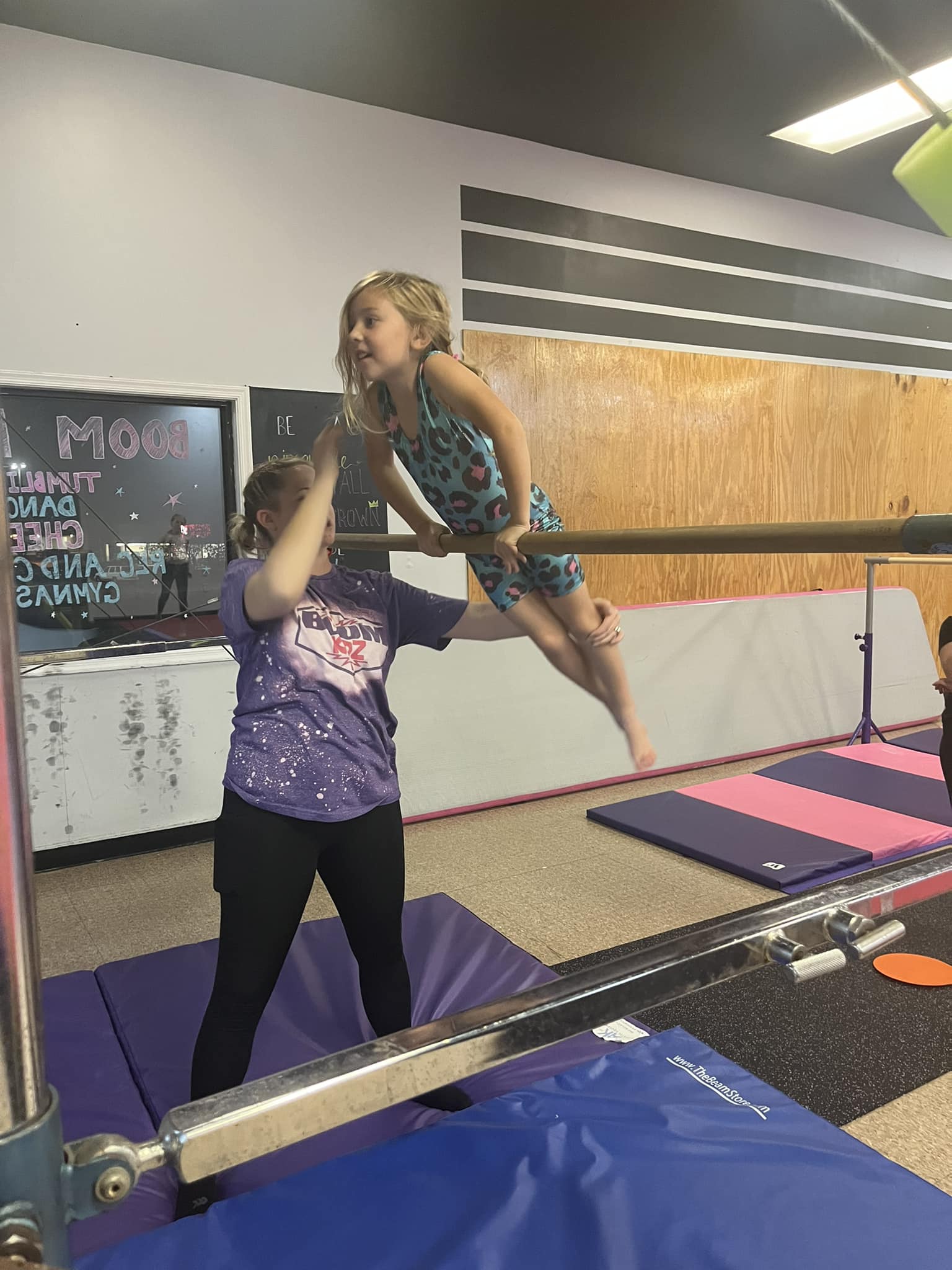 A young girl in a blue leotard practices on the uneven bars, supported by an instructor in a purple t-shirt. They are in a gymnastics facility with mats on the floor and equipment around. The instructor is helping the girl maintain her balance.