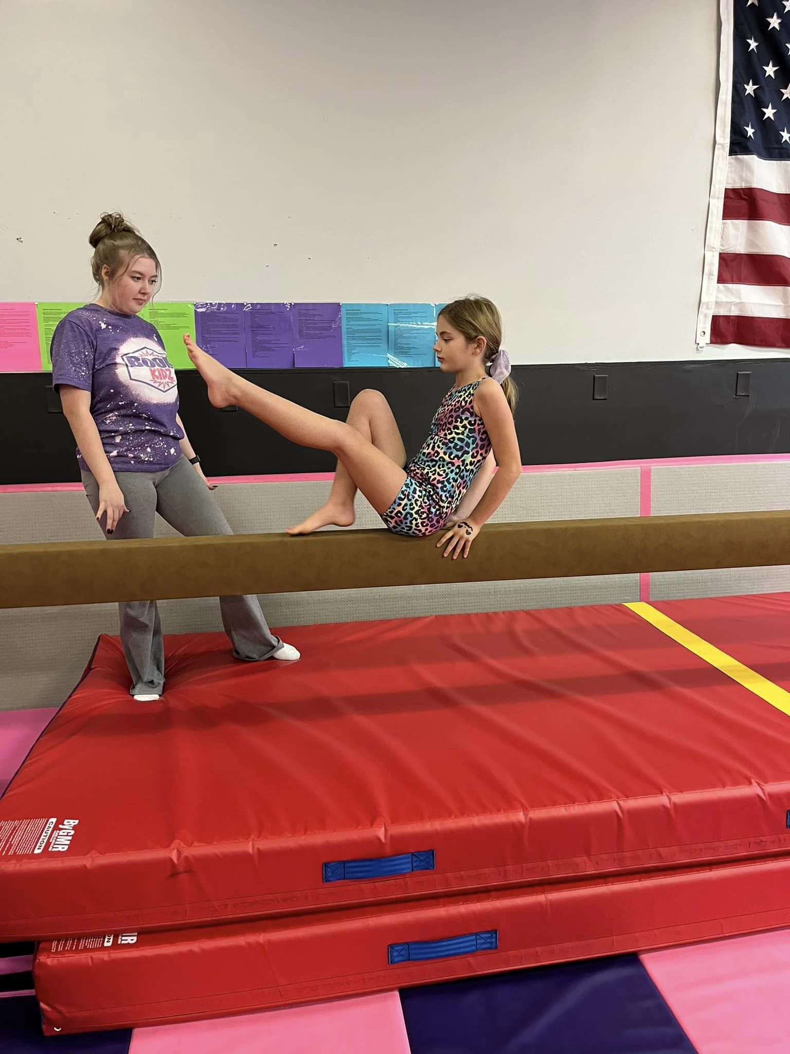 Two girls are in a gymnastics facility. One girl is sitting on a balance beam, extending her right leg. The other girl, standing on the mat below in a purple t-shirt, holds the extended leg. The background includes a U.S. flag and colorful papers on the wall.