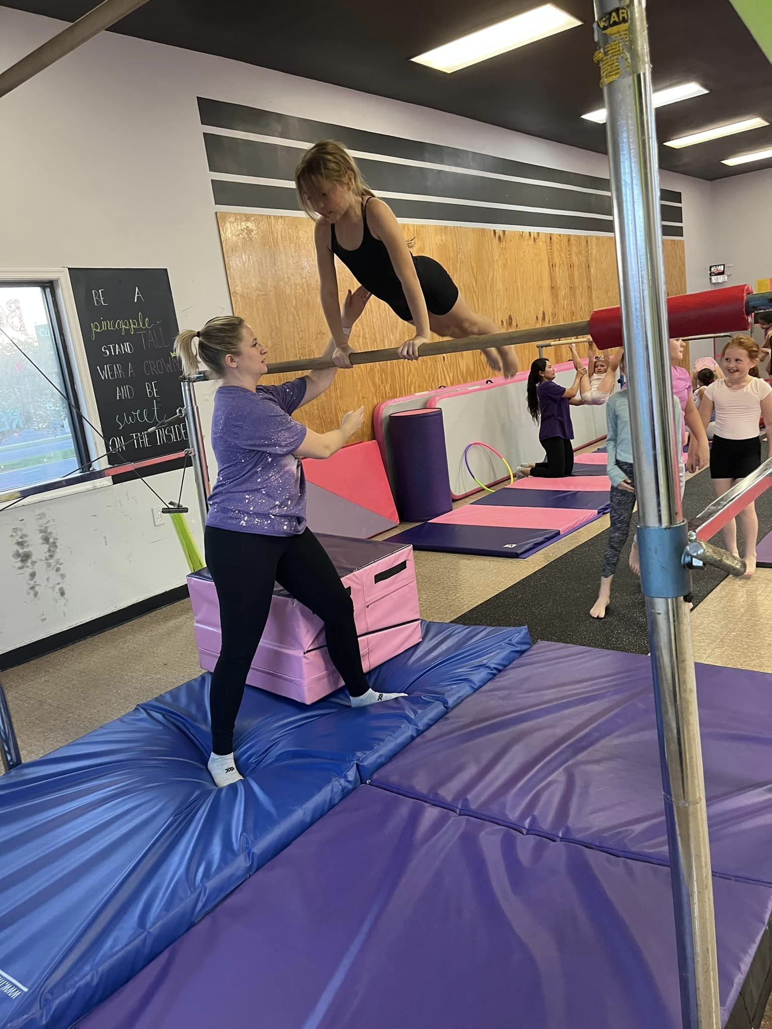 A young girl balances on a gymnastics horizontal bar while a coach stands on a padded block, assisting her. Other children and gym equipment are visible in the background. The gym has bright, colorful mats and a chalkboard with motivational writing on the wall.