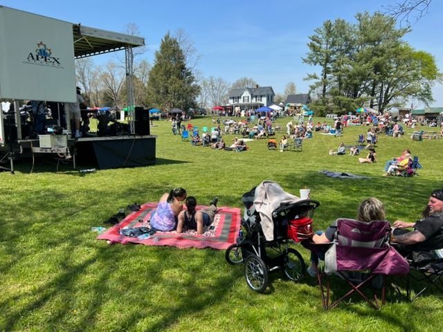 People gathered for an outdoor event in a sunny park with a stage on the left. Attendees sit on blankets and chairs, with some using strollers. Trees and a house are visible in the background, creating a relaxed, community atmosphere.
