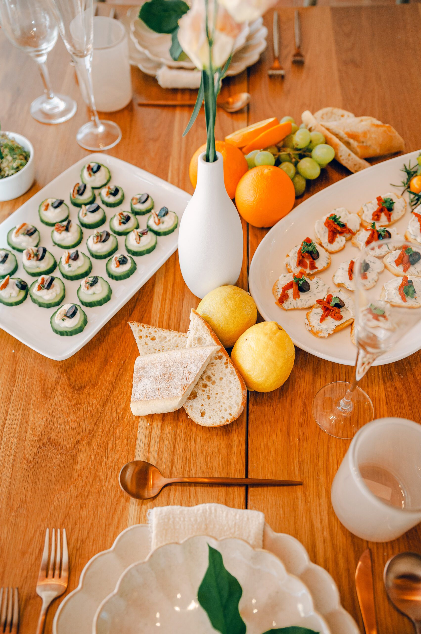 A wooden table set with an assortment of finger foods including cucumber bites on a white platter, lemon slices, bread, citrus fruits, and bruschetta. A white vase with a single flower and elegant glassware and plates complete the setup.