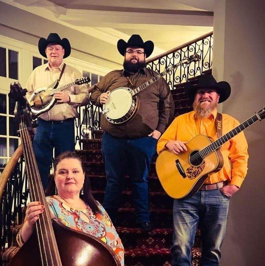 Four musicians stand on a staircase, posing with their instruments. A woman in front holds a double bass, while three men behind her hold a mandolin, a banjo, and an acoustic guitar. They are all wearing cowboy hats and casual clothing, embodying the lively spirit of bluegrass.