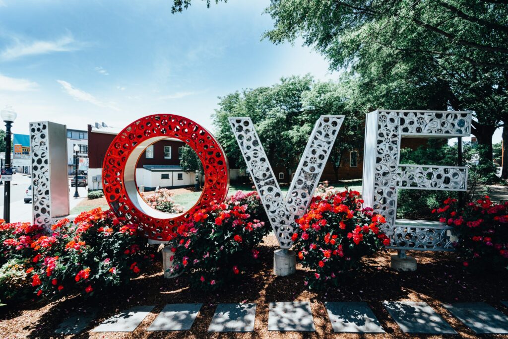 A large sculpture spelling "LOVE" stands amidst vibrant red flowers and greenery, flanked by colorful murals. The letters are artistically crafted, with the "O" featuring cut-out shapes. Trees canopy the scene under a bright blue sky, and buildings are visible in the background.