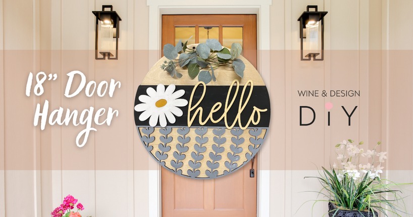 Front wooden door decorated with a round floral sign that says "hello" and greenery. The sign is black, gold, gray, and white, measuring 18 inches in diameter. Wall-mounted lanterns flank the door. Text on the image reads "18" Door Hanger" and "Wine & Design DIY.