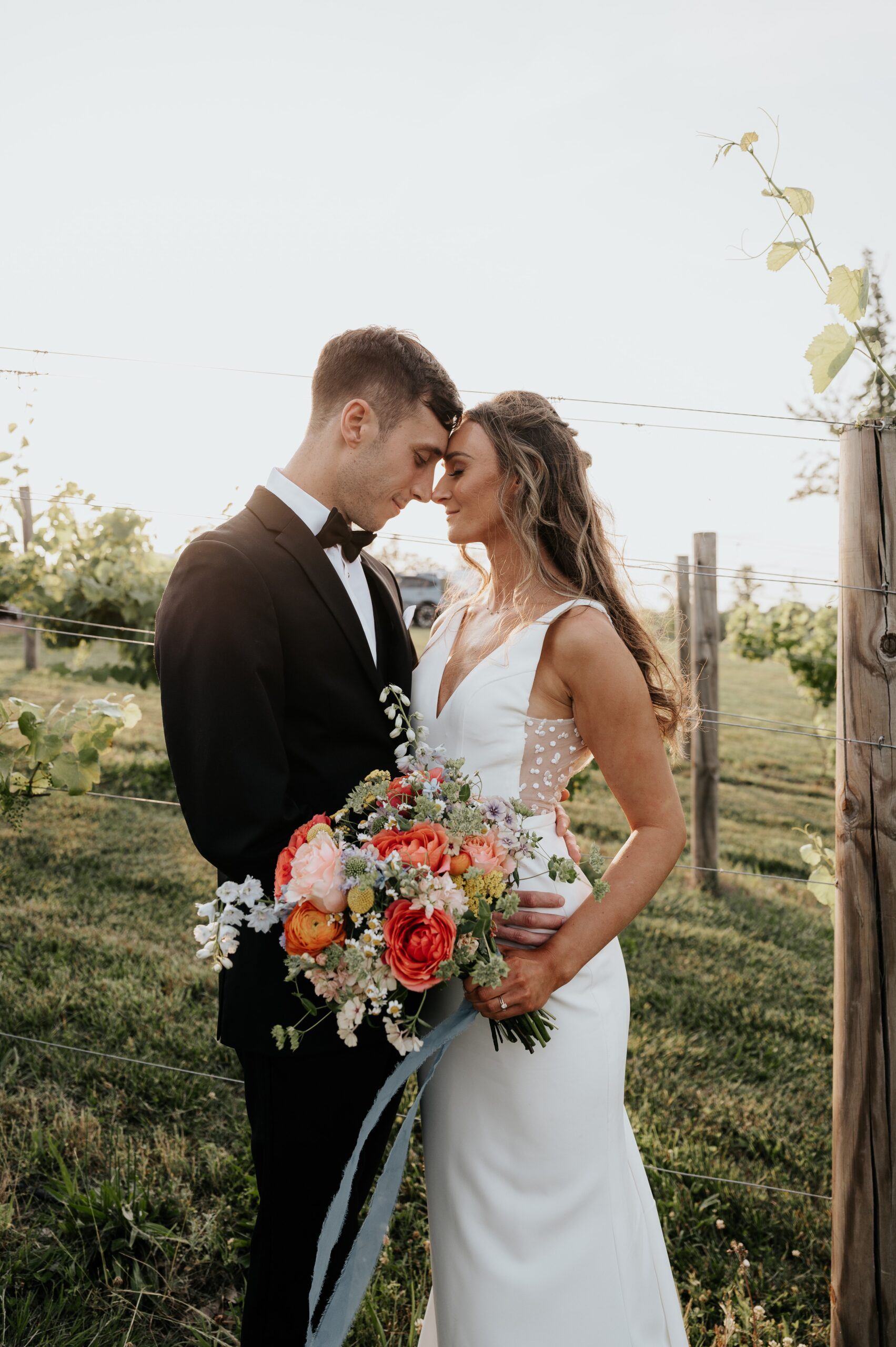 A bride and groom stand close together in an outdoor setting with a grassy background. The groom wears a black tuxedo while the bride wears a white dress and holds a vibrant bouquet of flowers. Both have their heads gently touching, looking content and happy.