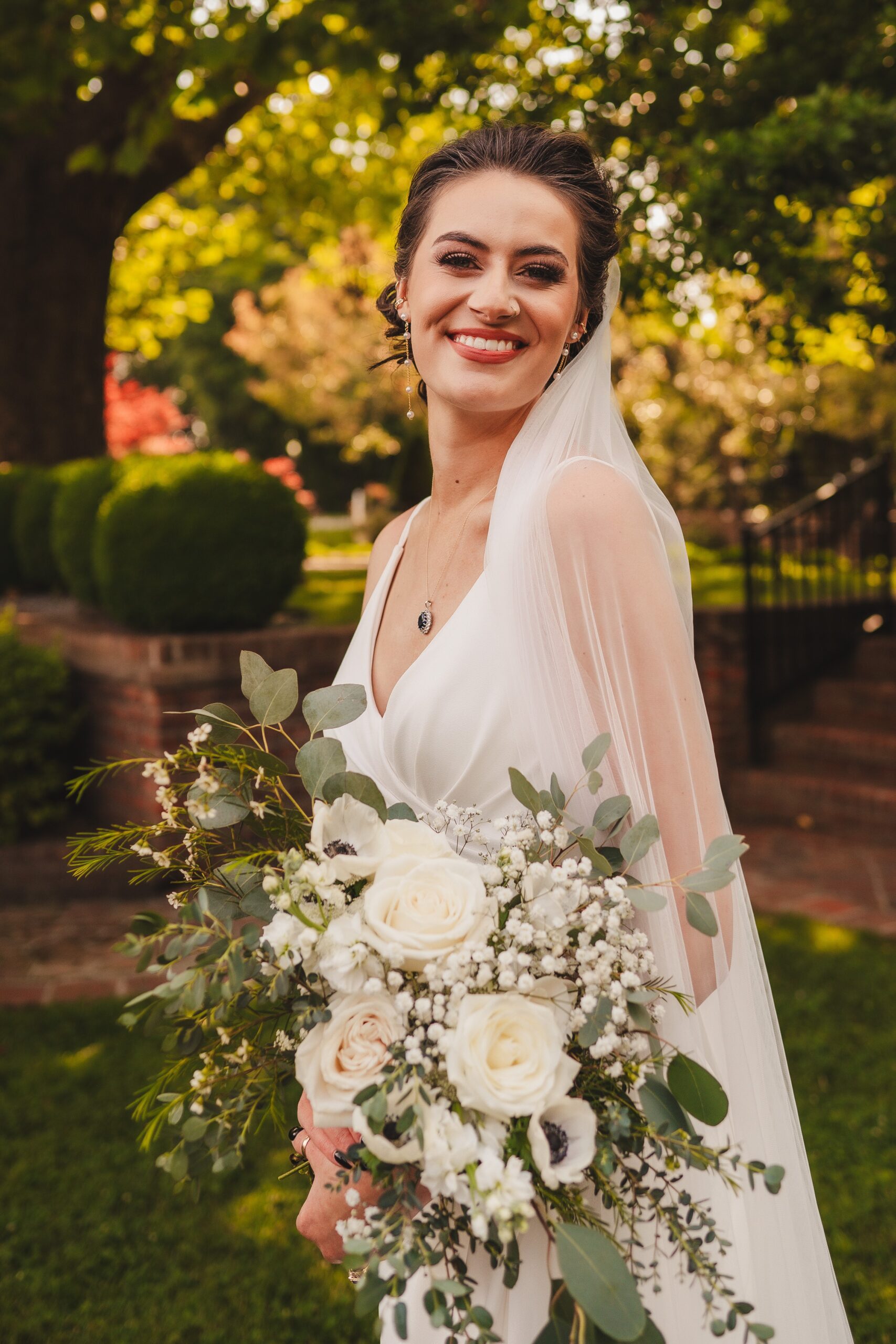 A bride with dark hair, wearing a white dress and veil, smiles warmly while holding a lush bouquet of white roses and greenery. She stands outdoors in a garden setting, with vibrant green trees and a sunlit background.