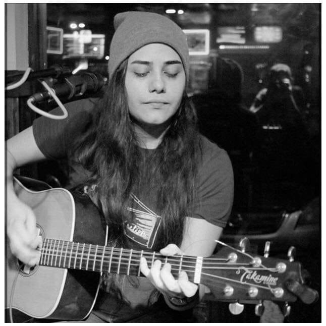 A black-and-white photo of a person with long hair wearing a beanie, strumming an acoustic guitar. They appear to be deep in concentration as they play. A microphone is positioned in front of them, and the background suggests a dimly lit room with reflections on glass.