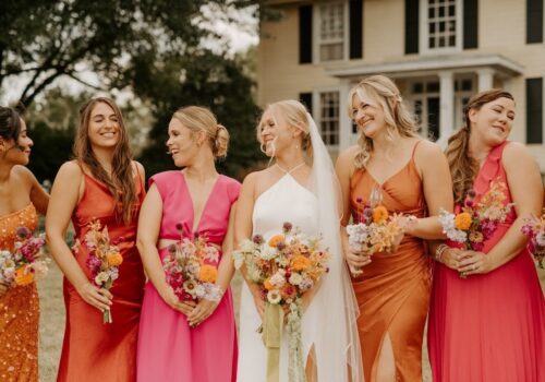 A bride in a white dress and veil stands smiling and holding a bouquet. She is surrounded by five bridesmaids wearing dresses in shades of pink, orange, and red. Each bridesmaid is also holding a colorful bouquet. A house and trees are visible in the background. Image