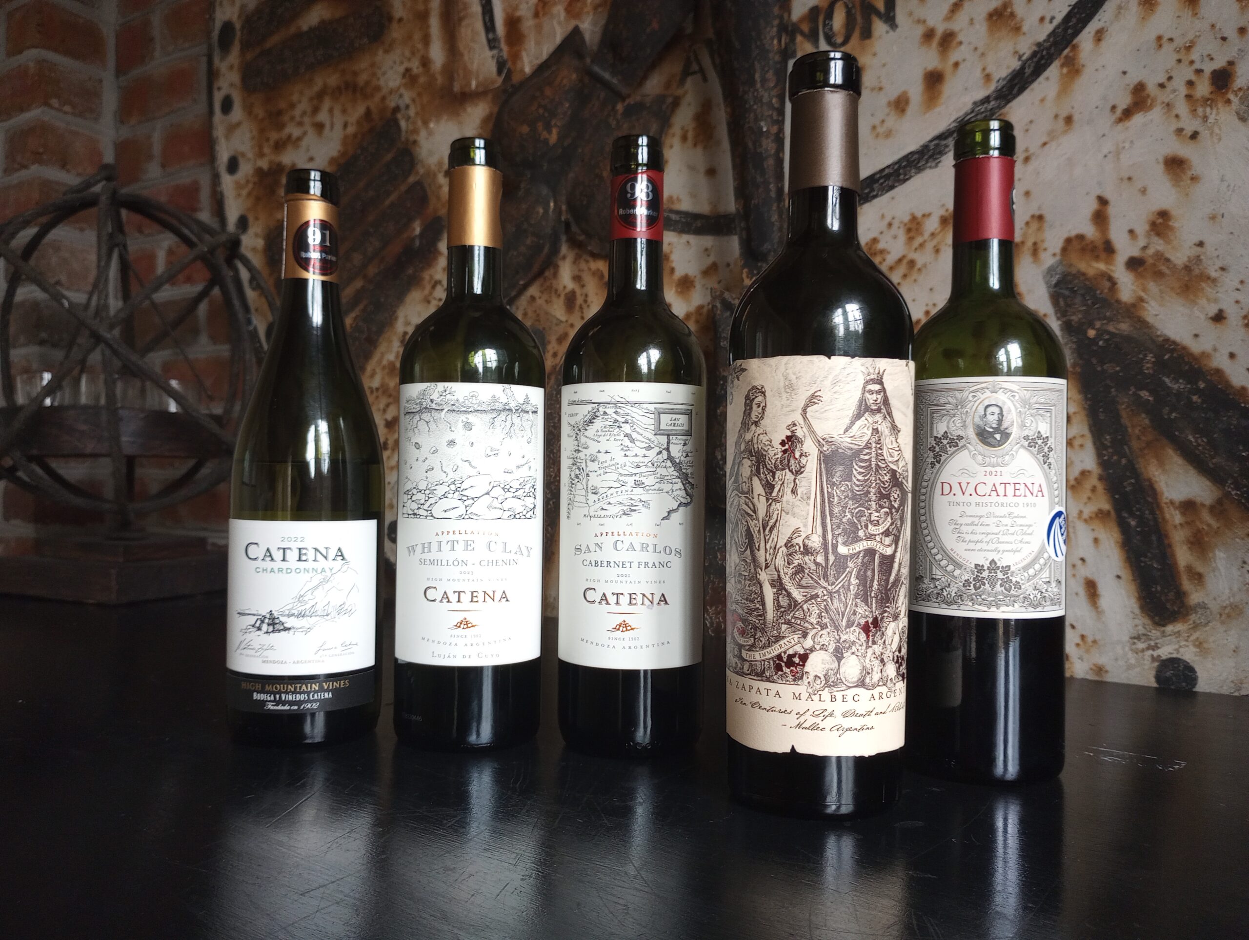 Five wine bottles stand in a row on a black table in front of a rustic, textured background. Each bottle features a distinct label with varying designs, including text and illustrations. Perfect for any wine dinner, the bottles differ in height and shape, showcasing a range of wine types.