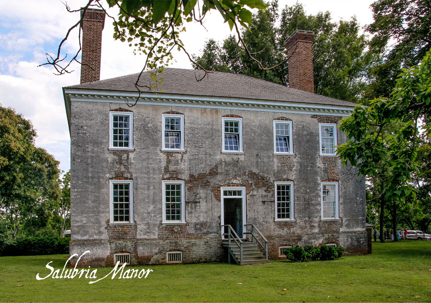 A historic, two-story house with weathered gray brick and tall chimneys stands surrounded by green grass and trees. The house has multiple large windows and a staircase leading to the front door. Text in the bottom left corner reads "Salubria Manor.