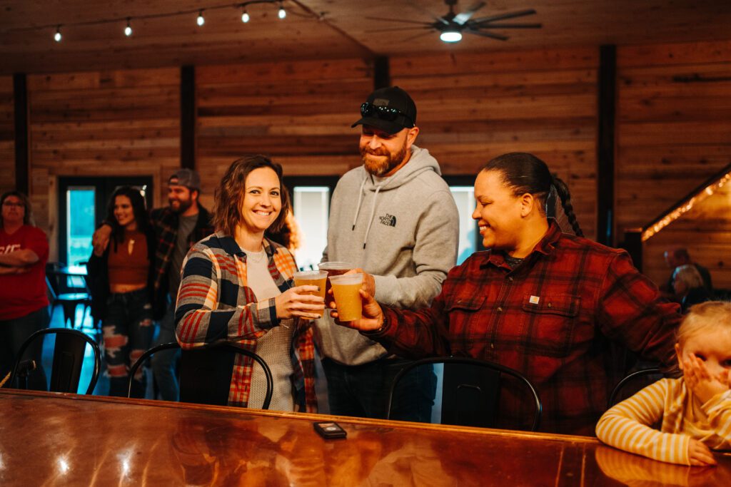 A group of people gathering indoors, with two of them at the bar holding and clinking glasses while smiling. The background features wood-paneled walls and several people interacting in a relaxed setting, as if they've just completed a local beverage trail.
