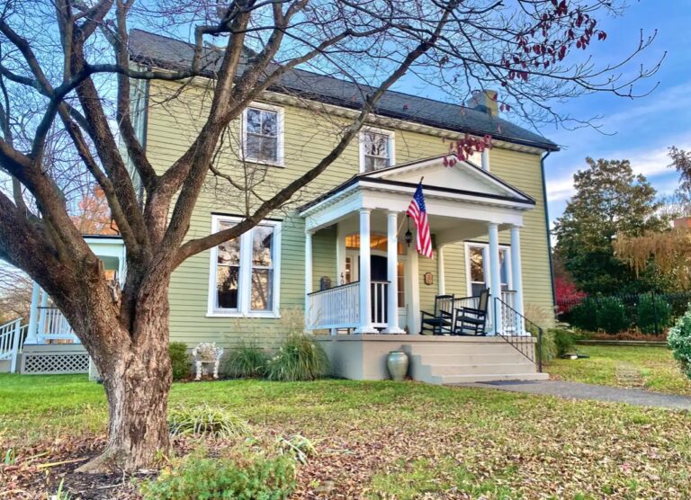 A charming two-story house with a green exterior and dark shutters serves as the perfect Spring Escape. It features a front porch with white columns, an American flag, and a rocking chair. A large leafless tree is in the foreground, surrounded by a well-maintained lawn and clear blue sky overhead.