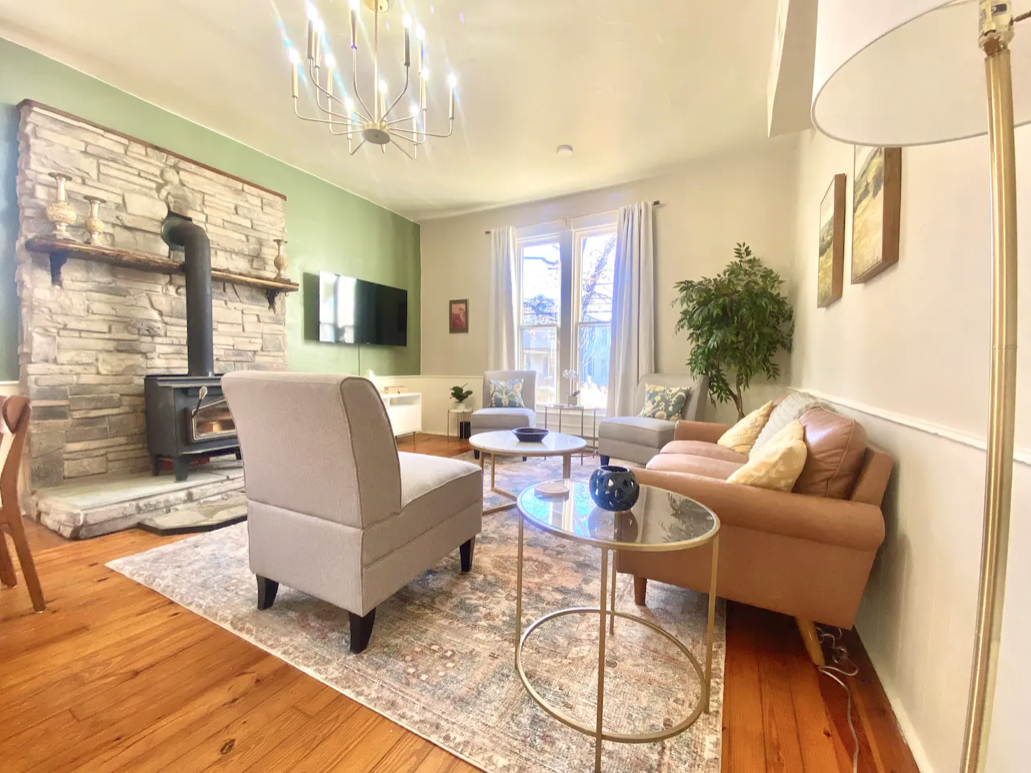 A cozy living room with a stone fireplace, modern wood-burning stove, and TV on a green accent wall. Reminiscent of Hill House charm, the space features a gray armchair, beige sofa with pillows, round glass coffee table, tall lamp, rug, and large plant near a window with curtains.