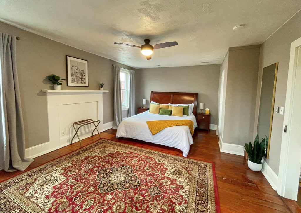 A spacious Hill House bedroom features a bed with a leather headboard, white bedding, and yellow pillows. A large ornate rug covers the wooden floor. The walls are painted light gray, and a ceiling fan hangs above the bed. A potted plant, bench, and wall décor complete the serene setting.
