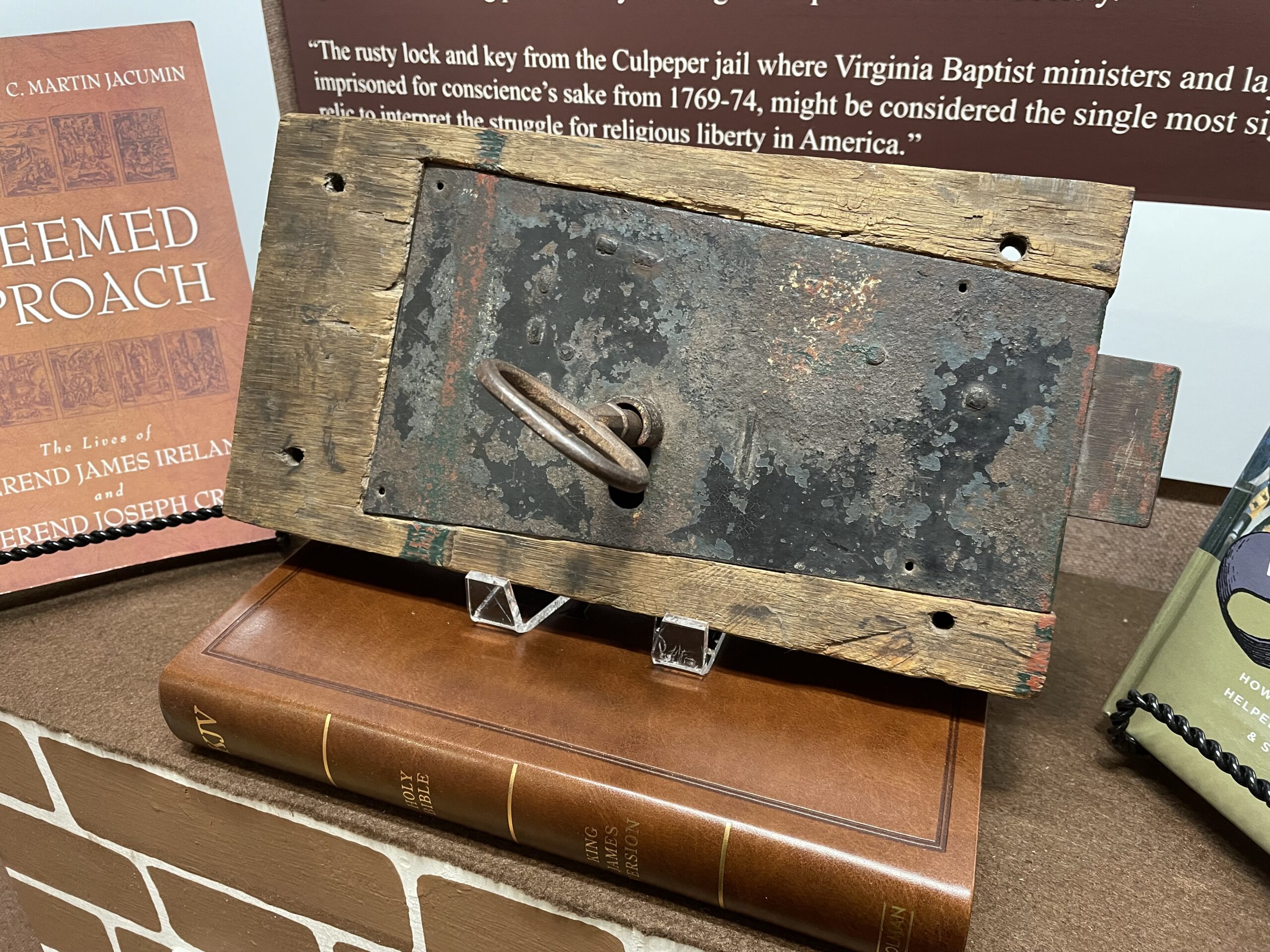A close-up of an old, rusted lock and key mounted on a wooden board displayed in the museum exhibit. The lock is weathered, indicating age. The display is set against a backdrop with historical text and books related to Baptist ministers and religious liberty in America.