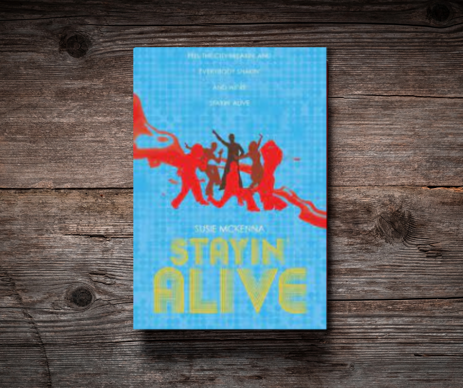 A book titled "Stayin Alive" by Susie McKenna rests on a wooden surface. The cover features vibrant silhouettes of people dancing in a celebratory pose against a blue background with red decorative elements.
