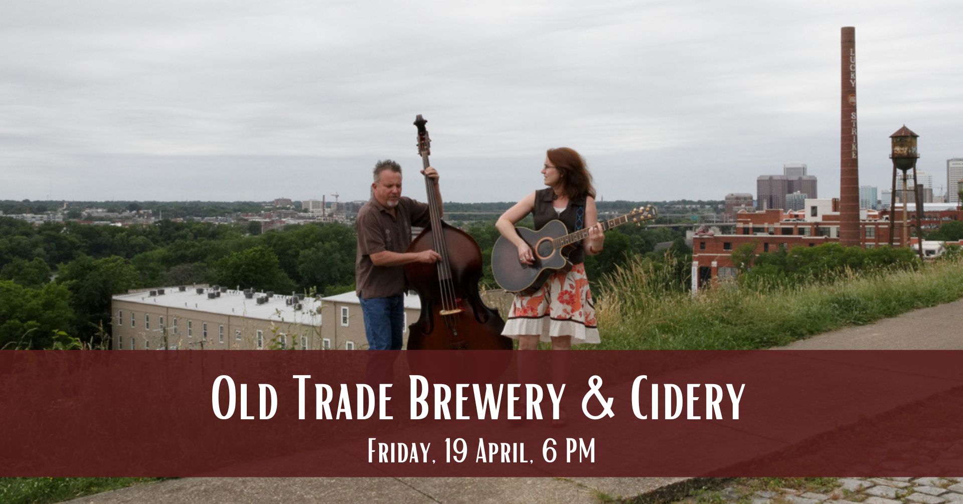 Two musicians perform with a double bass and acoustic guitar on a hill overlooking a cityscape. A red banner at the bottom reads, "Old Trade Brewery & Cidery, Friday, 19 April, 6 PM." The sky is overcast, and an industrial smokestack is visible in the background.