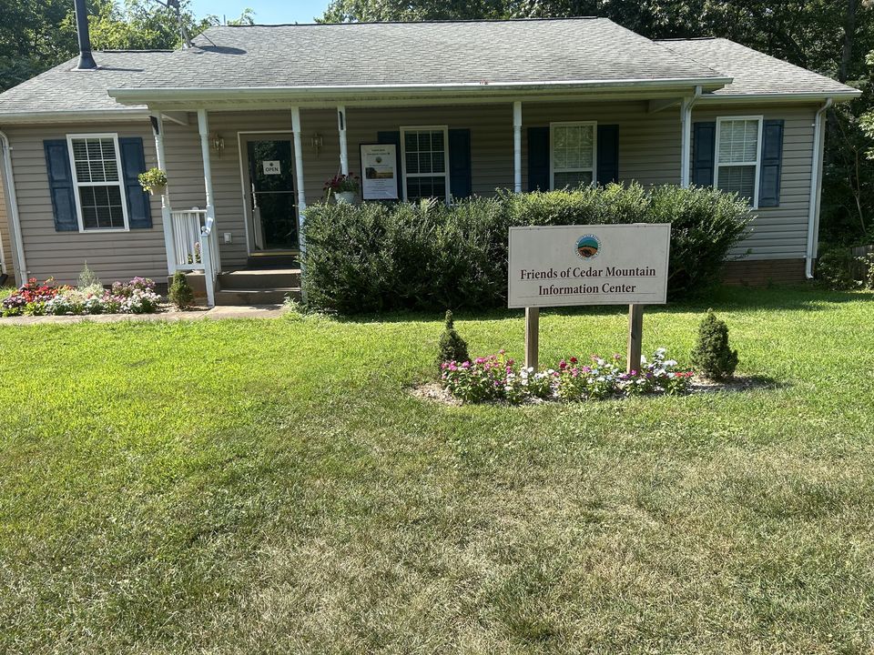 A small building with beige siding and blue shutters is surrounded by green grass and flowers. A sign in front reads, "Friends of Cedar Mountain Information Center." Bushes and a flower bed decorate the entrance, inviting guests to the upcoming Open House. Trees provide a serene backdrop.