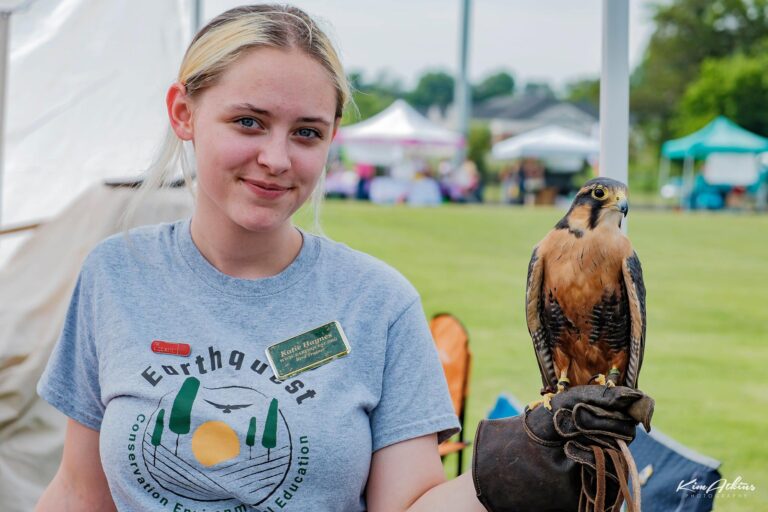A young woman wearing an EarthQuest t-shirt and a name tag stands at Free Fun in Culpeper, holding a falcon perched on her gloved hand. Tents and people are visible in the background on a grassy field.